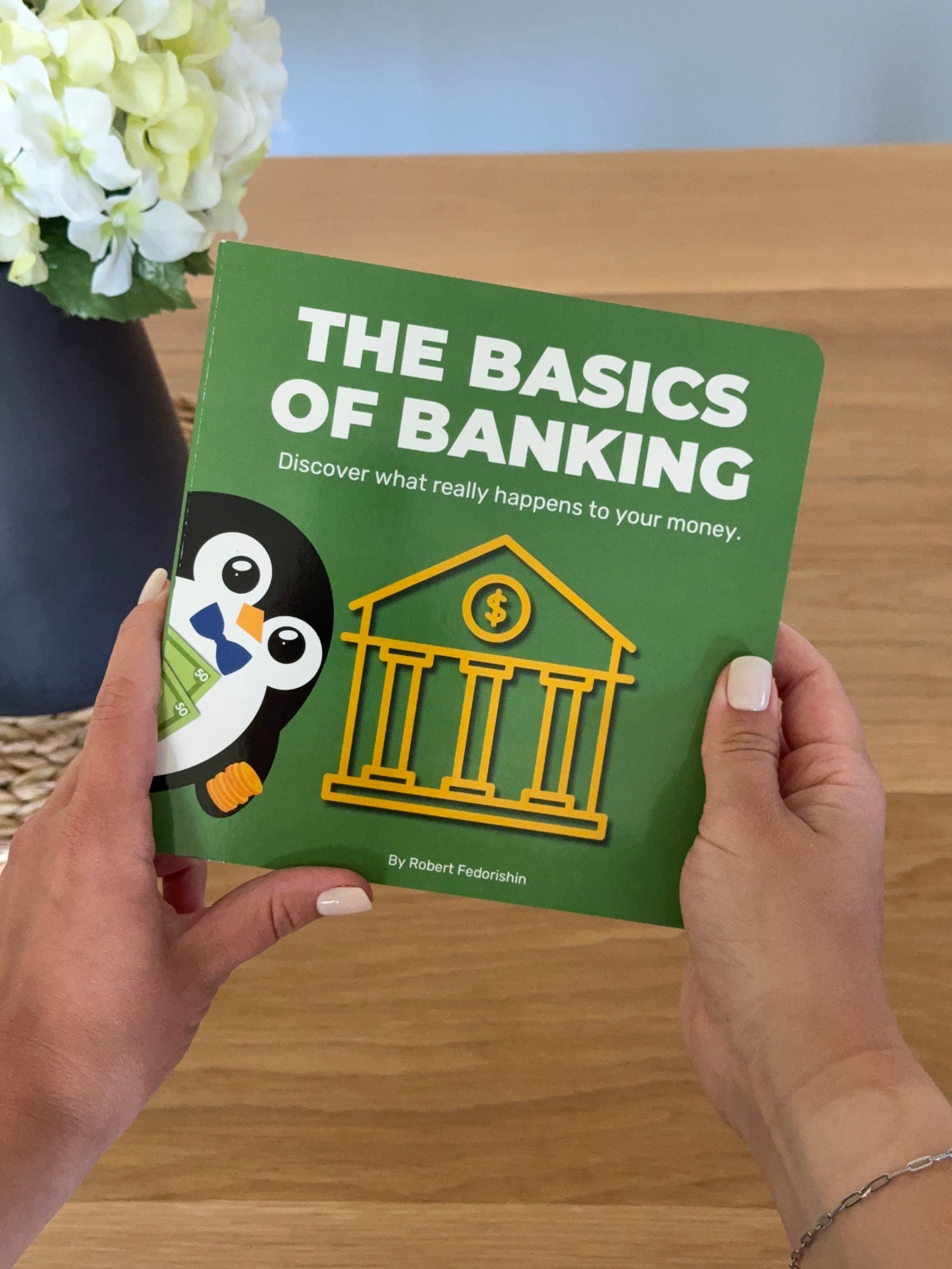 Person holding a children’s book about money and banking titled 'The Basics of Banking' with a penguin cartoon illustration.