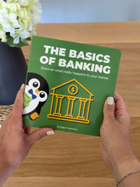 Person holding a children’s book about money and banking titled 'The Basics of Banking' with a penguin cartoon illustration.